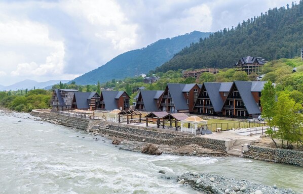 Un hotel a orillas del río con varias construcciones tipo cabaña, un muro de piedra de contención y montañas al fondo.