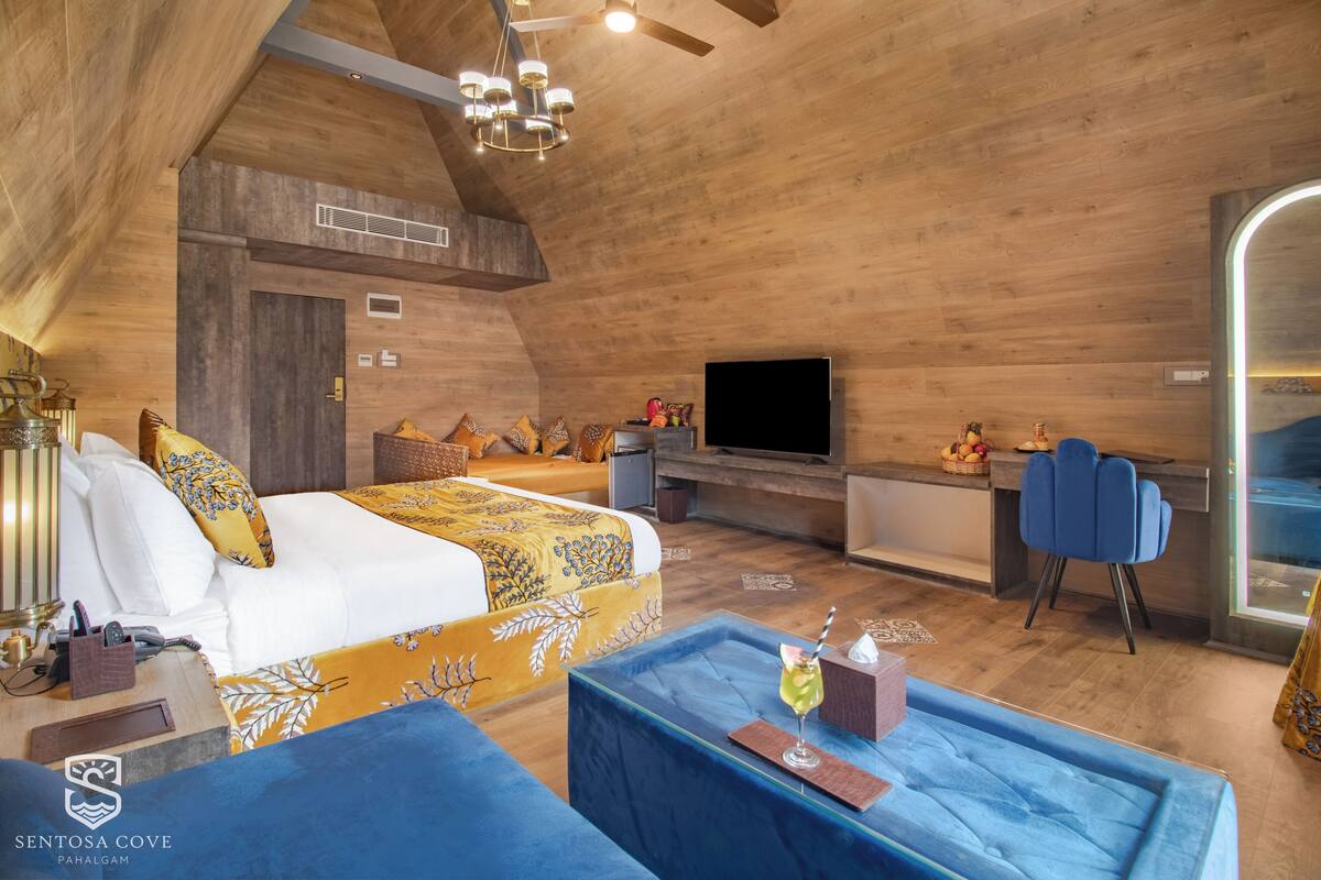 Un dormitorio con cama, mesita de noche, televisión y una zona de estar con sofá azul y una pequeña mesa.