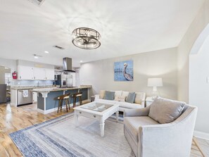 A modern kitchen with white cabinets, a central island with bar stools, a dining table, and a living area with a sofa and a lamp.