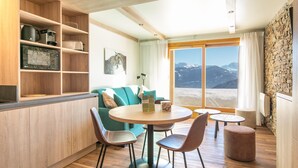 A modern room with a wooden floor, a round table with chairs, a sofa, a bookshelf, and a view of mountains through a large window. - Scenic Mountain Stay for Four (La Léchère)