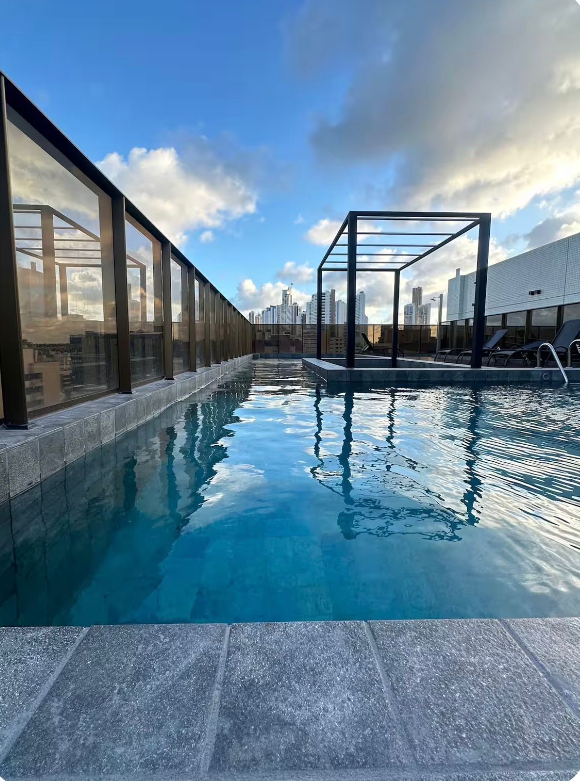 Una piscina infinita en la azotea con vistas a un horizonte urbano, una estructura de cristal y sillones de descanso.