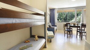 Ein Zimmer mit einem Etagenbett, einer Schlafcouch, einem Esstisch mit Stühlen und Blick auf Grün durch große Fenster.
