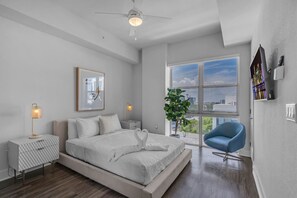 Ein modernes Schlafzimmer mit Bett, Nachttischlampen, einem blauen Sessel, einem Blumentopf und Blick auf die Stadt.