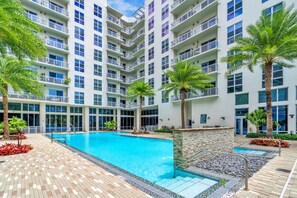 Ein mehrstöckiges Hotel mit einem zentral gelegenen Schwimmbecken, umgeben von Palmen und einer Ziegelterrasse.