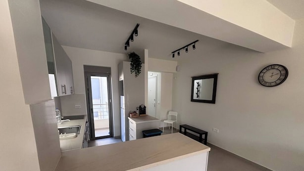 A modern kitchen with a central island, a dining area with a bench, a white chair, a clock on the wall, and a door leading to a balcony. - Vasili's Mini Villa in Nafplio With Shaded Parking (Nafplio)