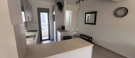 Una cocina moderna con una isla central, un área de comedor con un banco, una silla blanca, un reloj en la pared y una puerta que da a un balcón.