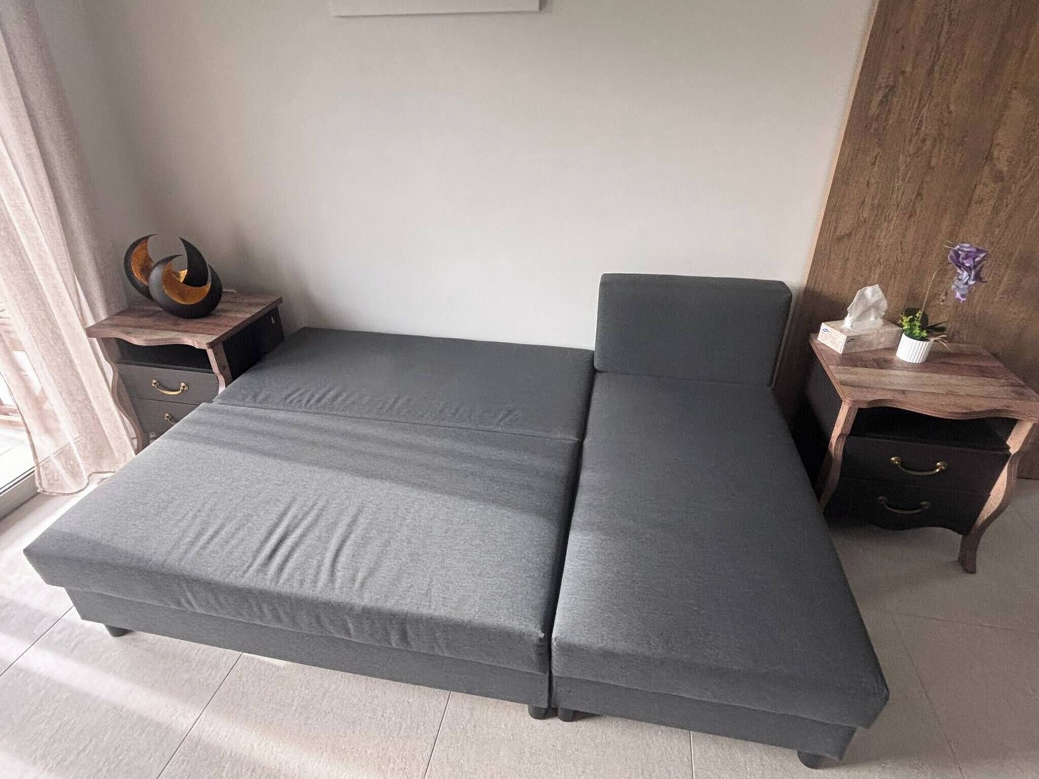 A modern room with a grey sofa bed, a small wooden side table with a decorative item, and a nightstand with a plant.