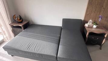 A modern room with a grey sofa bed, a small wooden side table with a decorative item, and a nightstand with a plant.