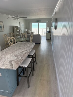 A kitchen with a marble countertop, wooden stools, a ceiling fan, and a view of the ocean through a large window. - Just Listed - Enjoy sunset every night with direct beach and pool access. (Fort Myers Beach)
