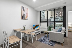 A modern living room with a dining table, chairs, a sofa, a TV, and a large window with a view of buildings. - Elevated 1-bed Retreat Near Crown (Melbourne)