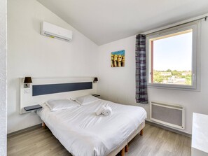Een netjes opgemaakt bed met hoofdbord, nachtkastje met lamp, wandairconditioner, radiator en raam met uitzicht op groen.
