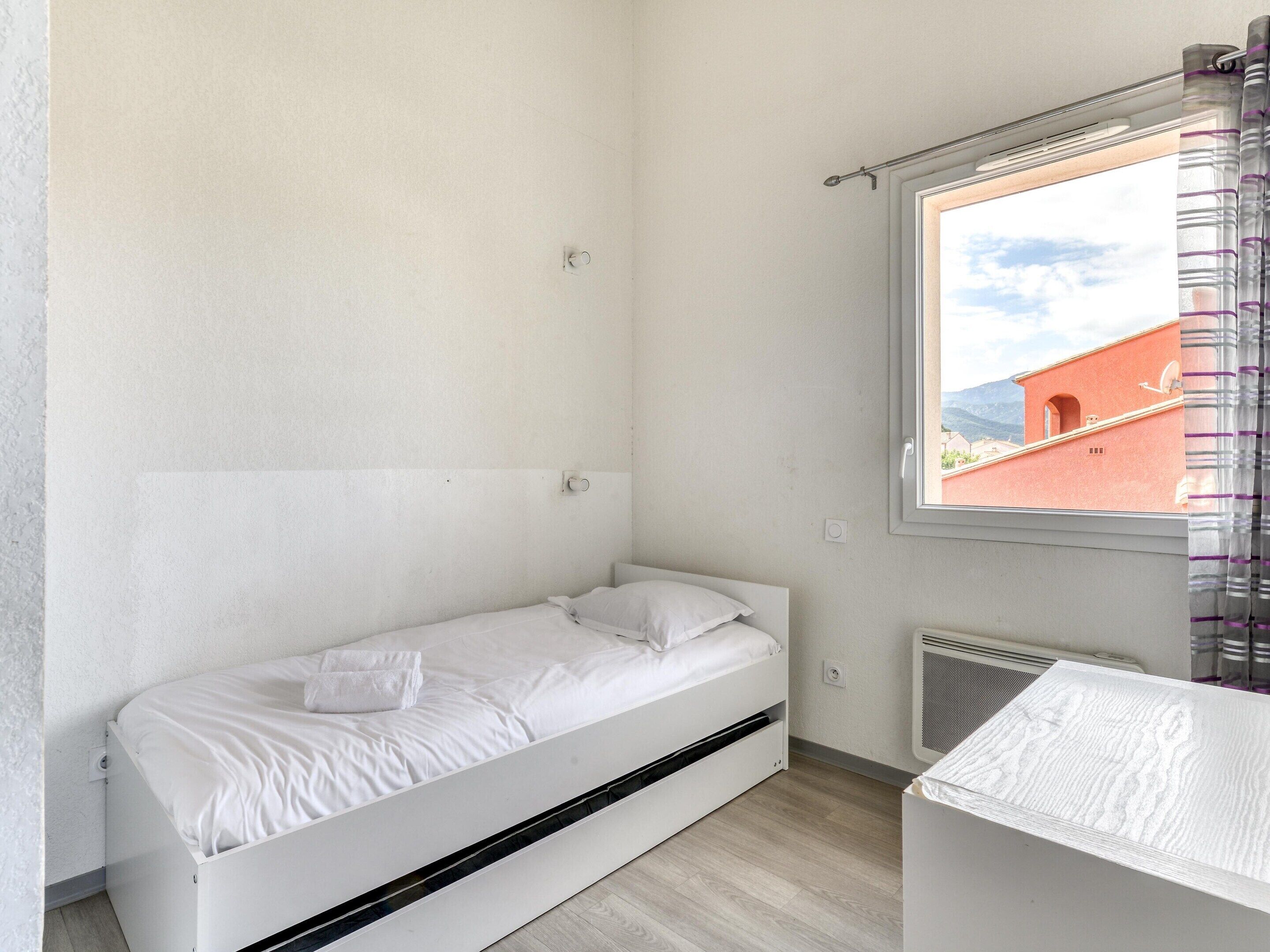 Habitación pequeña con cama individual, ventana, radiador y vista a un edificio.