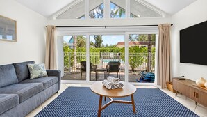 A living room with a grey sofa, a round wooden table, and a large sliding glass door leading to a balcony with a pool and greenery. - Beachfront Complex! Heated Pool~Pool View! Updated (Holmes Beach)