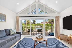 A living room with a grey sofa, a round wooden table, and a large sliding glass door leading to a balcony with a pool and greenery.