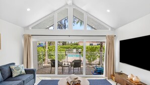 A modern living room with a large sliding glass door leading to an outdoor patio area, a flat-screen TV mounted on the wall, and a sofa with a patterned cushion. - Beachfront Complex! Heated Pool~Pool View! Updated (Holmes Beach)
