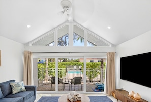 A modern living room with a large sliding glass door leading to an outdoor patio area, a flat-screen TV mounted on the wall, and a sofa with a patterned cushion.