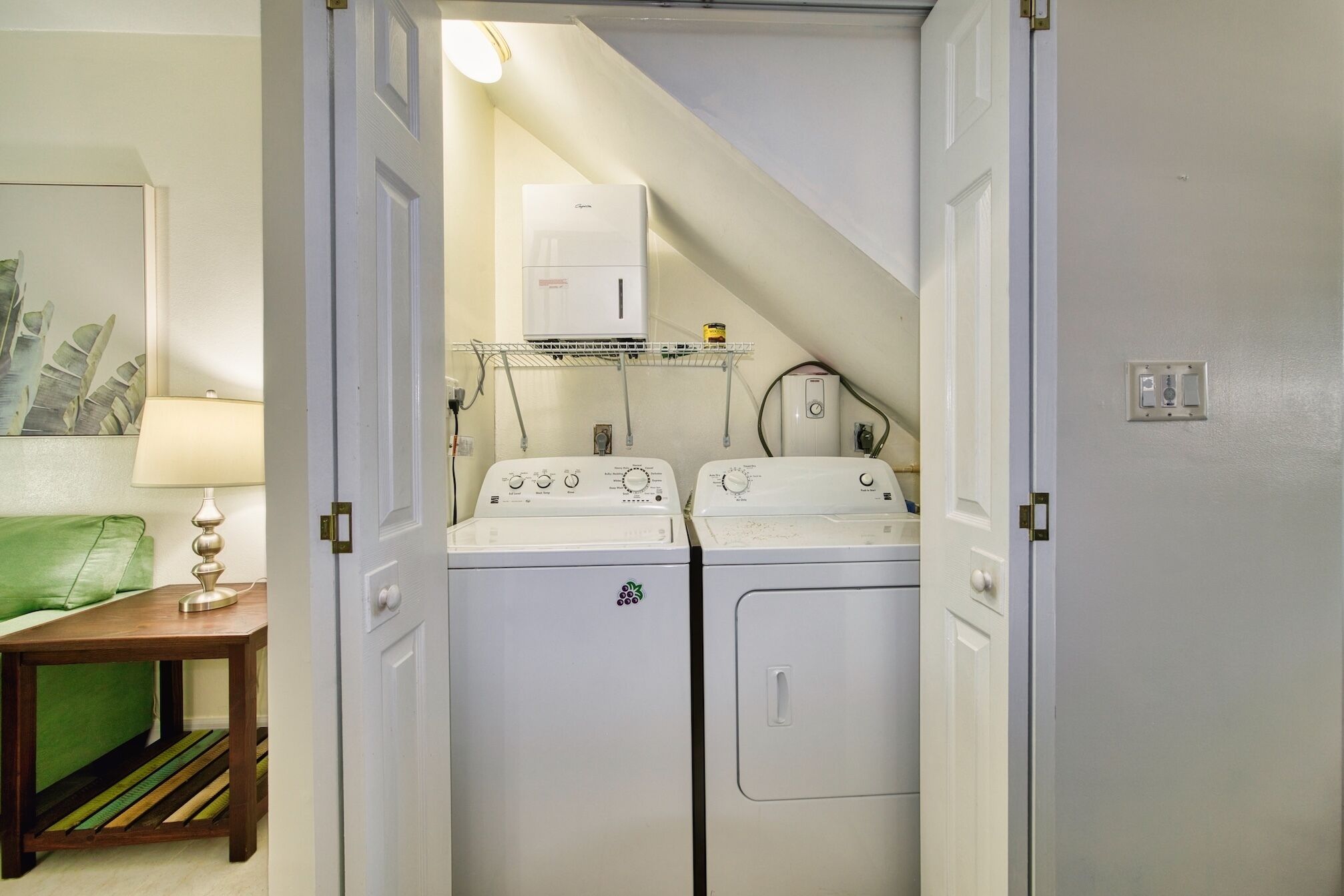 A laundry room with two white washing machines, a shelf with laundry supplies, and a green couch in the adjacent room.
