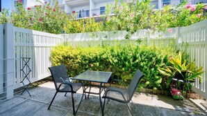 A patio with a table and chairs, surrounded by lush greenery and a white fence. - 2BR/2BA Peaceful Retreat, Patio, Walk to Beach (West Bay)