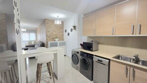 Eine moderne Küche mit eingebauten Geräten, ein Essbereich mit weißem Tisch und Stühlen sowie ein Wohnbereich mit einer Couch und einer Wanddekoration.
