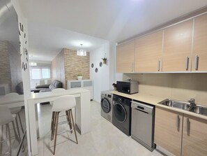 Una cocina moderna con electrodomésticos integrados, una zona de comedor con una mesa y sillas blancas, y una sala de estar con sofá y un detalle decorativo en la pared.