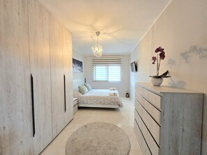 Un dormitorio con cama, cómoda, mesita de noche, candelabro, ventana con persianas y un cuadro en la pared.