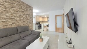 Ein Wohnzimmer mit grauem Sofa, einem weißen Couchtisch, einem an der Wand montierten Fernseher und einer Küche im Hintergrund.
