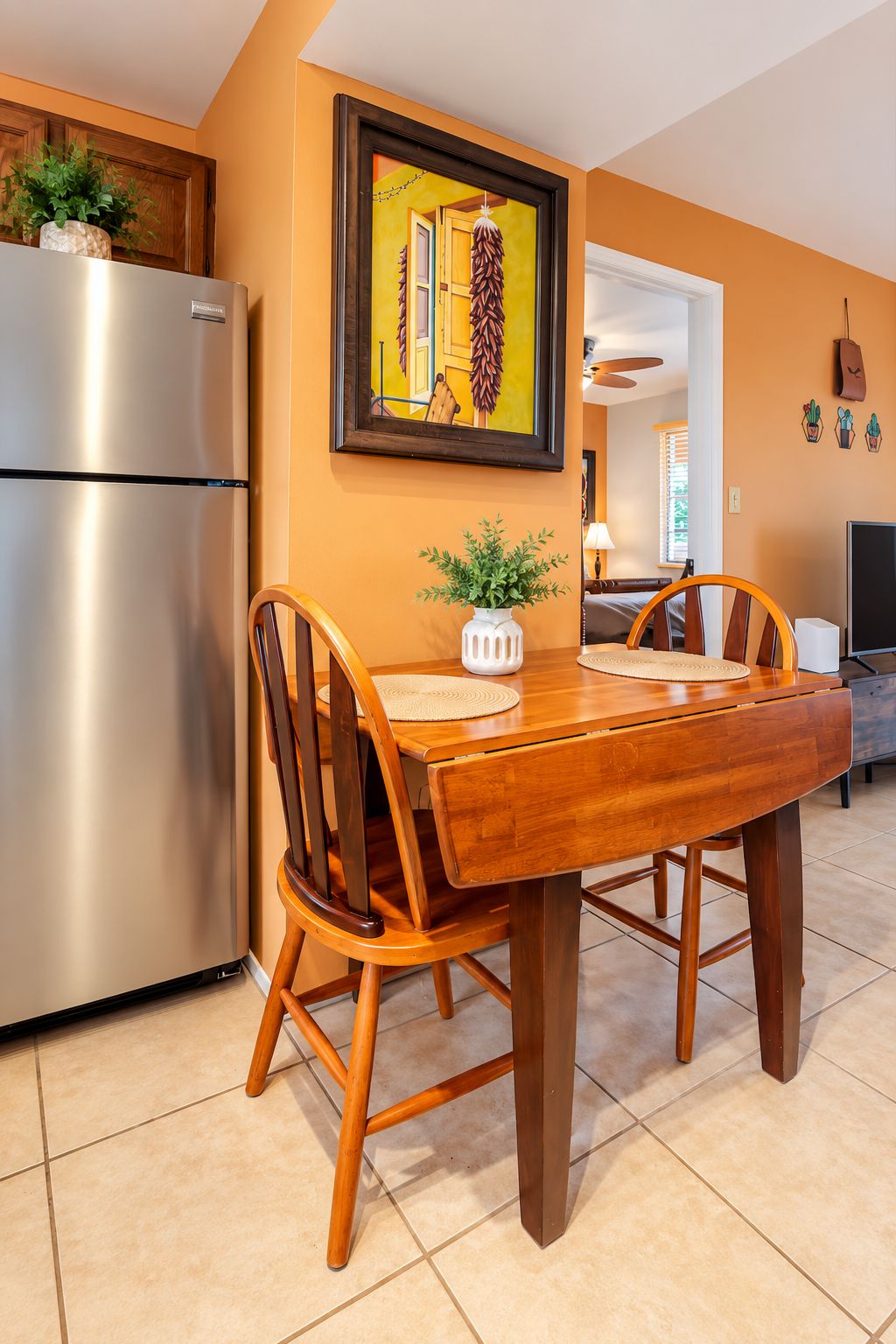 Una cocina moderna con refrigerador de acero inoxidable, mesa y sillas de madera, un cuadro en la pared y plantas en macetas.