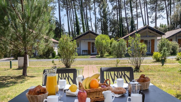 Tavolo da pranzo all'aperto apparecchiato con prodotti per la colazione, circondato da alberi e un edificio sullo sfondo.