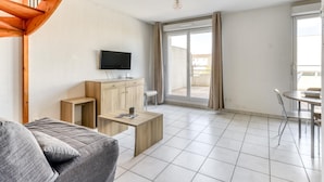 A modern living room with a sofa, a TV, a wooden cabinet, and a dining area with a table and chairs. - Pool View Apartment for Six (Le Grau-Du-Roi)