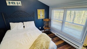 Un dormitorio con cama, una silla, una ventana con persianas y un cuadro en la pared.
