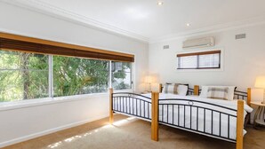 A bedroom with a bed, two bedside tables, a window with blinds, an air conditioning unit, and a view of greenery outside. - Metawise Stylish 2-bedroom Home With Easy CBD Ac (Point Clare)