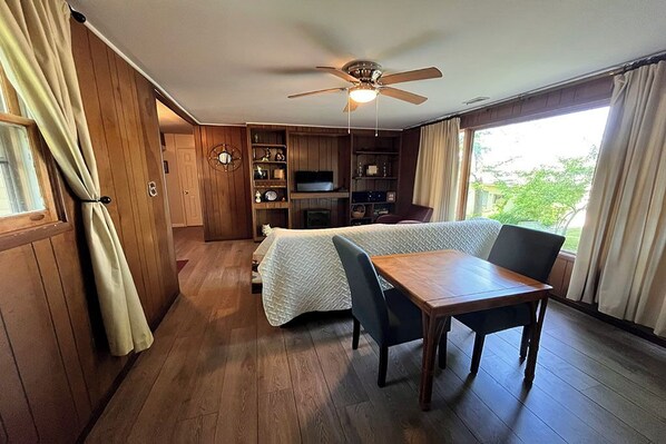 Una habitación con piso de madera, ventilador de techo, cama con colcha blanca, mesa de comedor con sillas y televisor sobre una estantería.