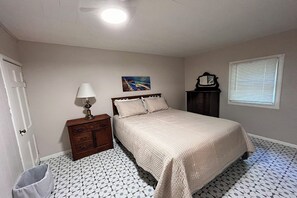 A bedroom with a bed, a nightstand, a lamp, a painting, a clock, and a window with blinds.
