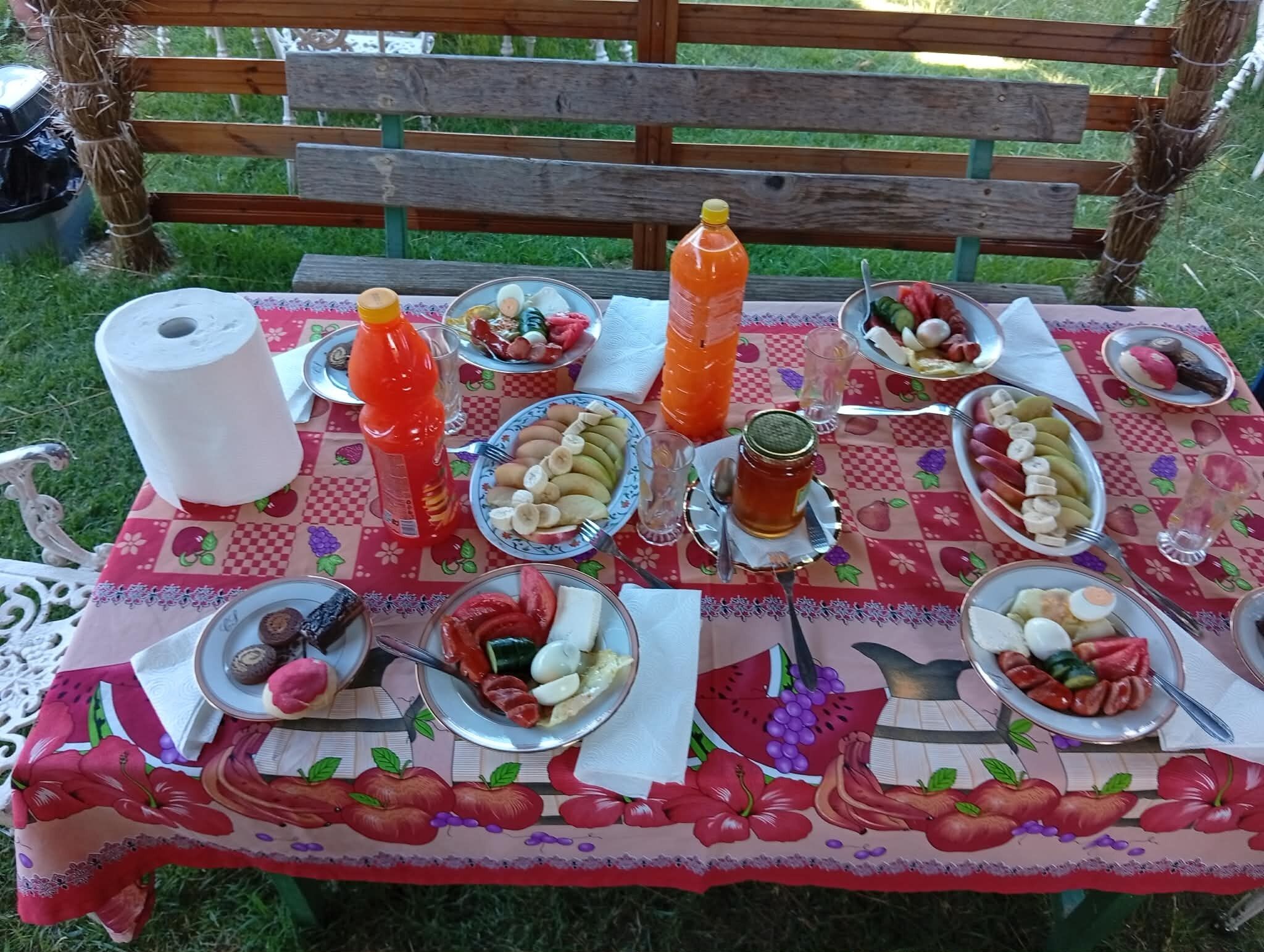 Montaje para picnic con una mesa cubierta por un mantel estampado con motivos de frutas, diferentes alimentos, una botella de jugo de naranja y un rollo de toallas de papel.