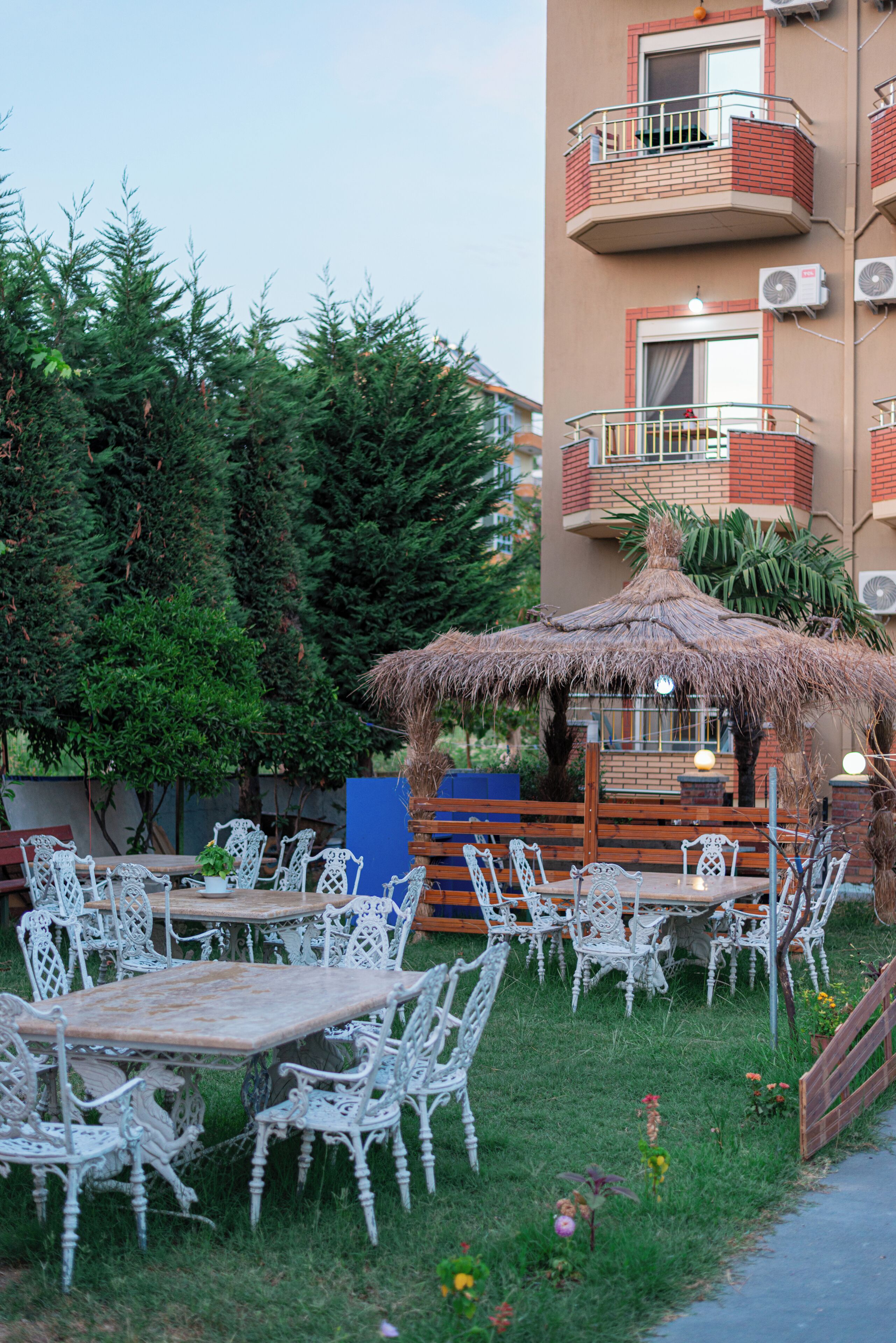 An outdoor seating area with white wicker chairs and tables, a thatched-roof structure, and a building with balconies and air conditioning units in the background.