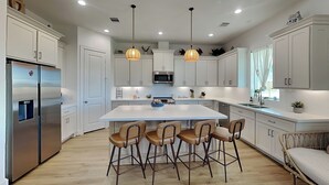 A modern kitchen with white cabinets, a stainless steel refrigerator, a central island with bar stools, pendant lighting, and a microwave. - Buckin Beach House (Galveston)