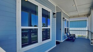 A covered porch with a railing, large windows, and a seating area with chairs. - Buckin Beach House (Galveston)