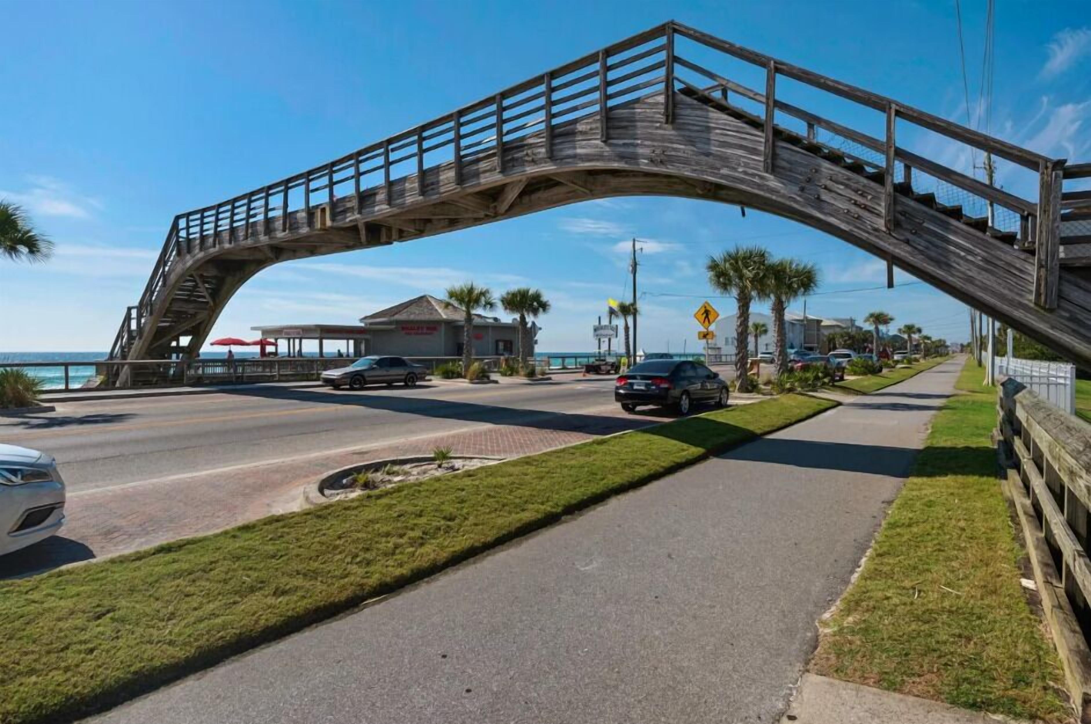 Un puente peatonal de madera sobre una carretera con coches, aceras y un edificio con toldo rojo al fondo.