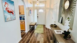 A hallway with wooden flooring, a bench, a dining table with a vase of flowers, and a wall-mounted mirror.