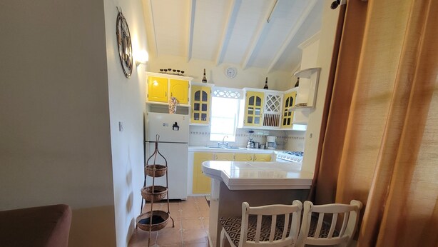 A kitchen with yellow cabinets, a white refrigerator, a sink, and a stove. There are two white chairs and a small table.