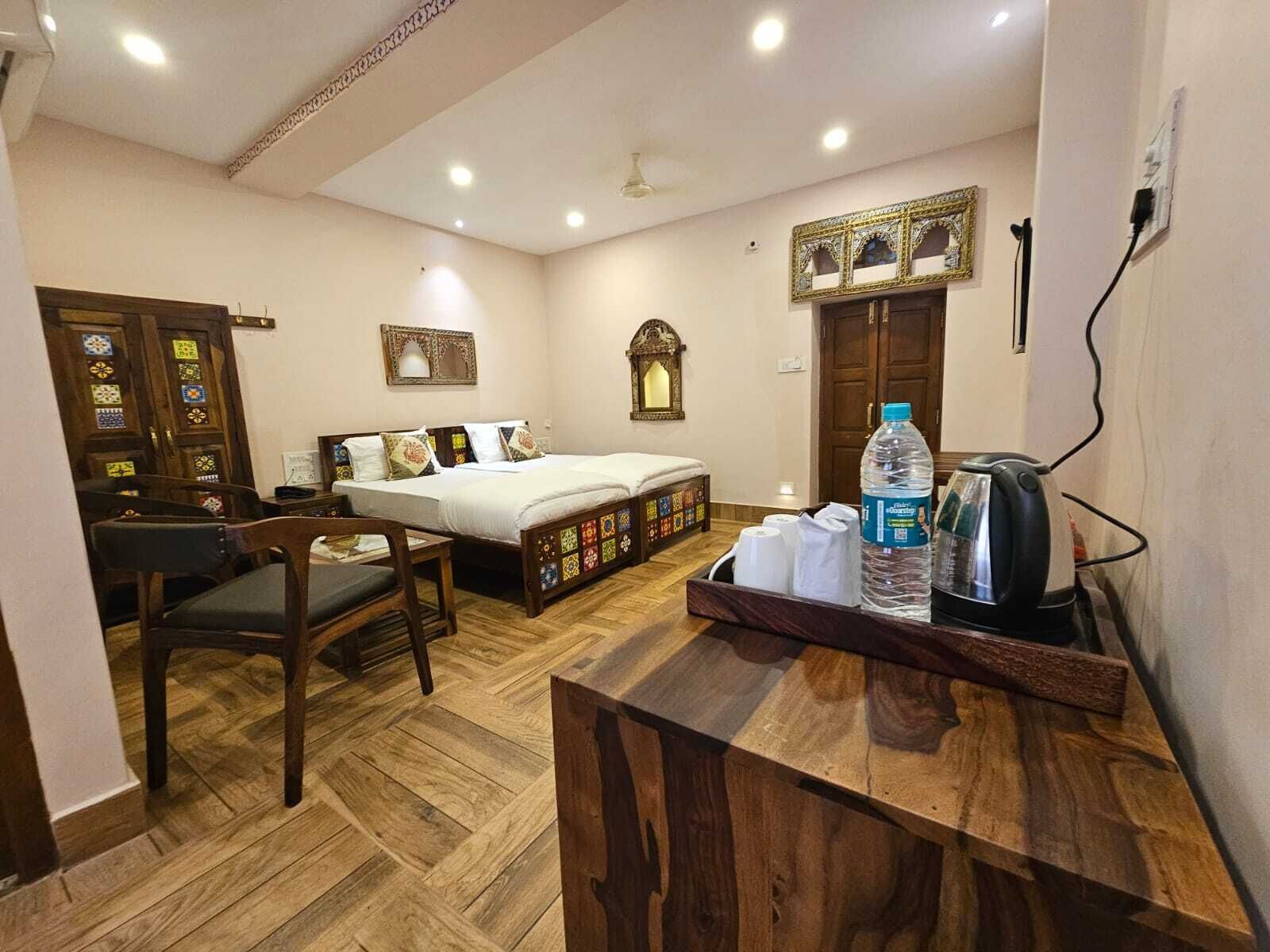 Habitación de hotel con cama, escritorio, silla, botella de agua, tetera y un espejo decorativo.