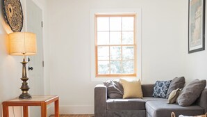A living room with a wooden floor, a gray sofa, a small wooden table, a lamp, a window, and a ceiling light. - Harriss-James Carriage House Unit B, 0.3 Mi from Historic Downtown (Wilmington)
