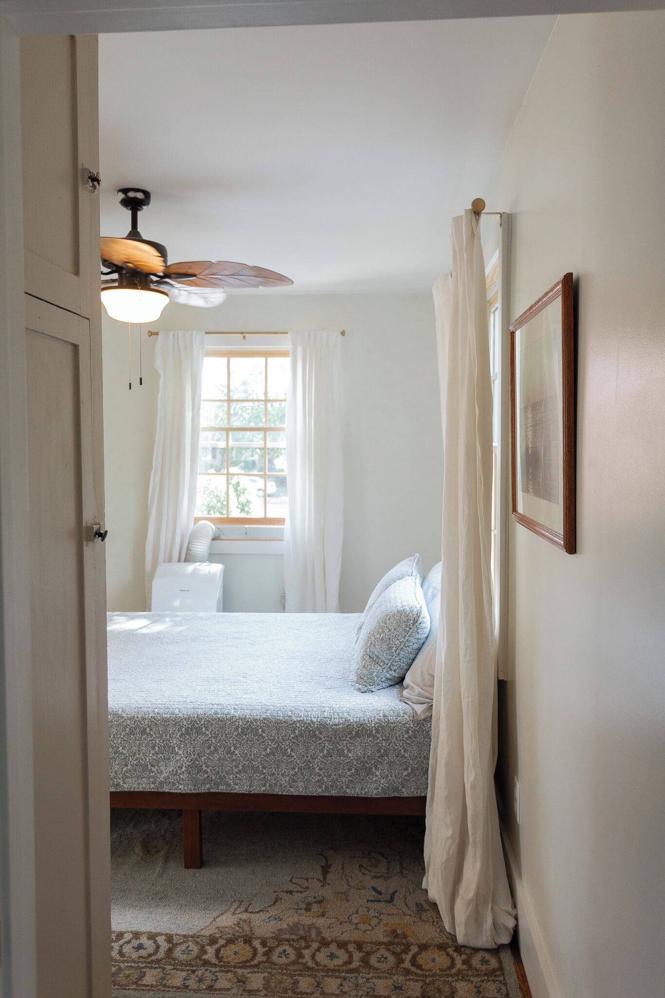 Une chambre à coucher avec un lit, une fenêtre avec des rideaux, un ventilateur de plafond et un tableau encadré au mur.