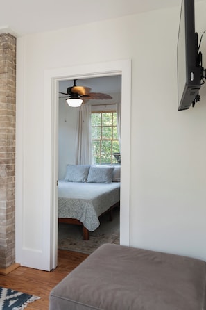 Ein Schlafzimmer mit einem Bett, einem Deckenventilator, einem Fenster mit Vorhängen und einem an der Wand montierten Fernseher.