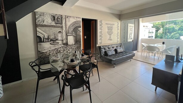 A dining area with a glass table, chairs, and a sofa. There is a staircase on the left and a balcony with a view outside. - LK Santo Antônio 4 (Belo Horizonte)