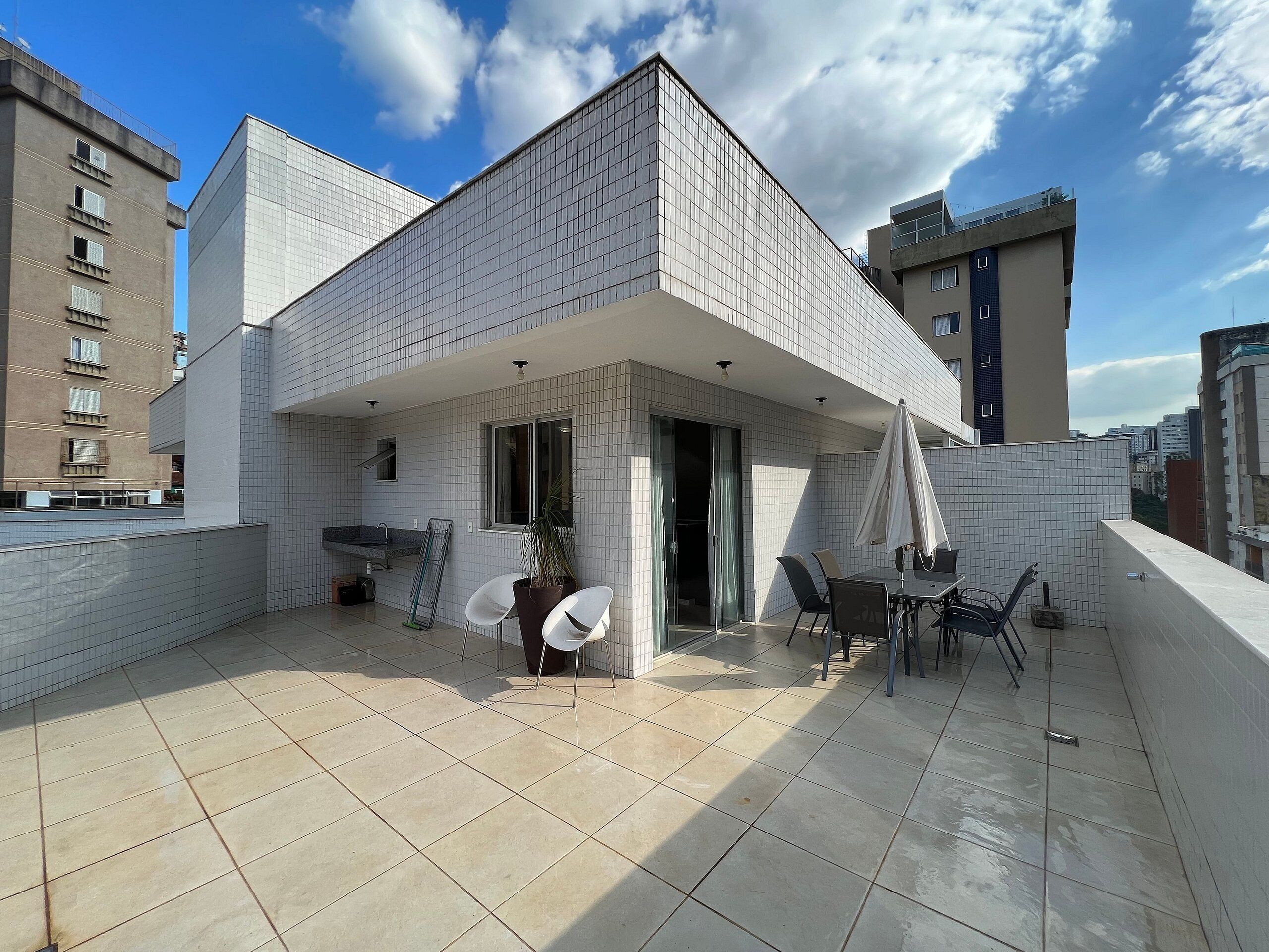 A modern building with a tiled exterior, a balcony with chairs and a table, and a view of surrounding buildings.