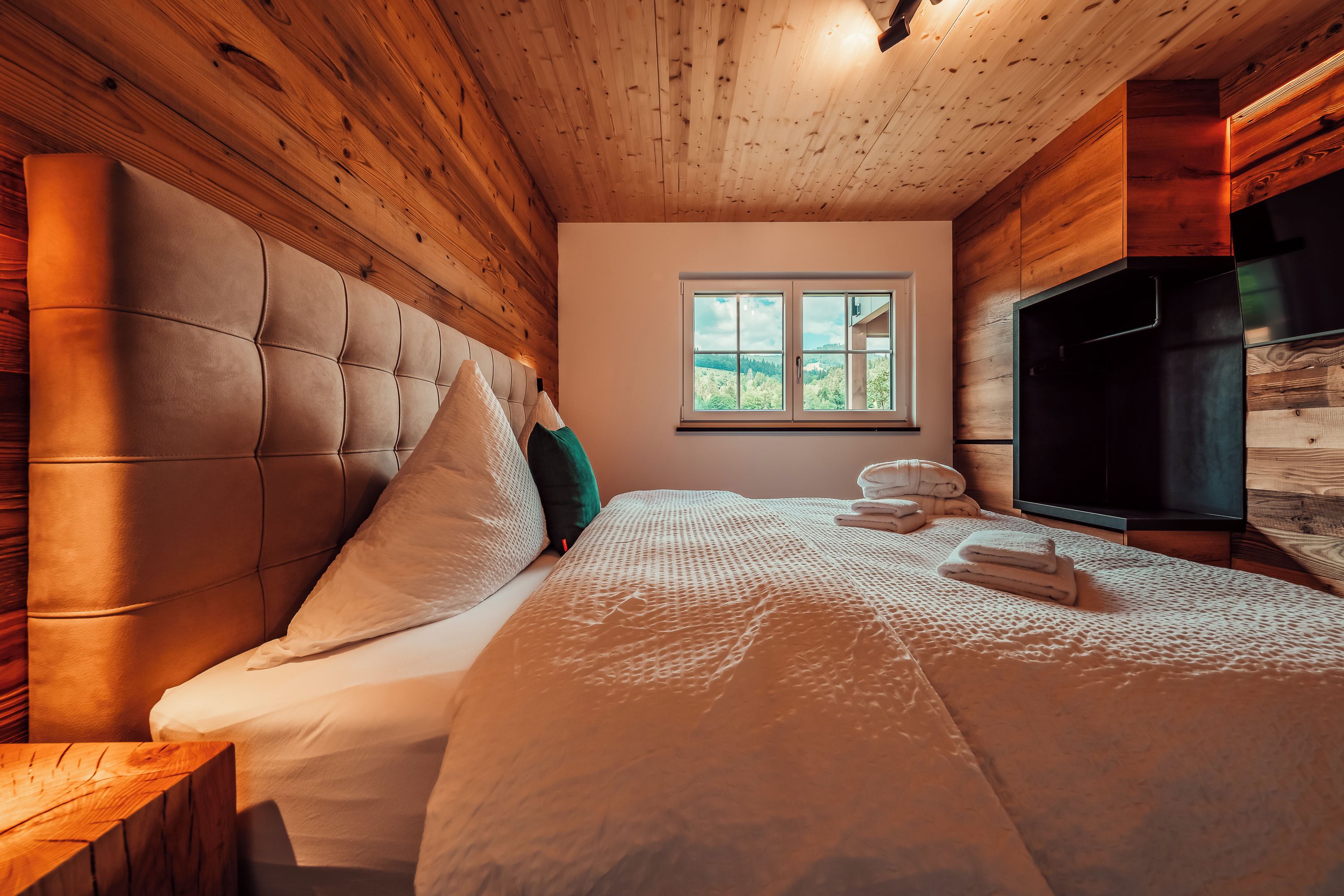 Une chambre avec un plafond en bois, un grand lit avec de nombreux oreillers, une fenêtre donnant sur un beau paysage et un téléviseur.