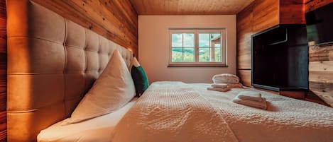 A bedroom with a wooden ceiling, a large bed with multiple pillows, a window with a view, and a television.