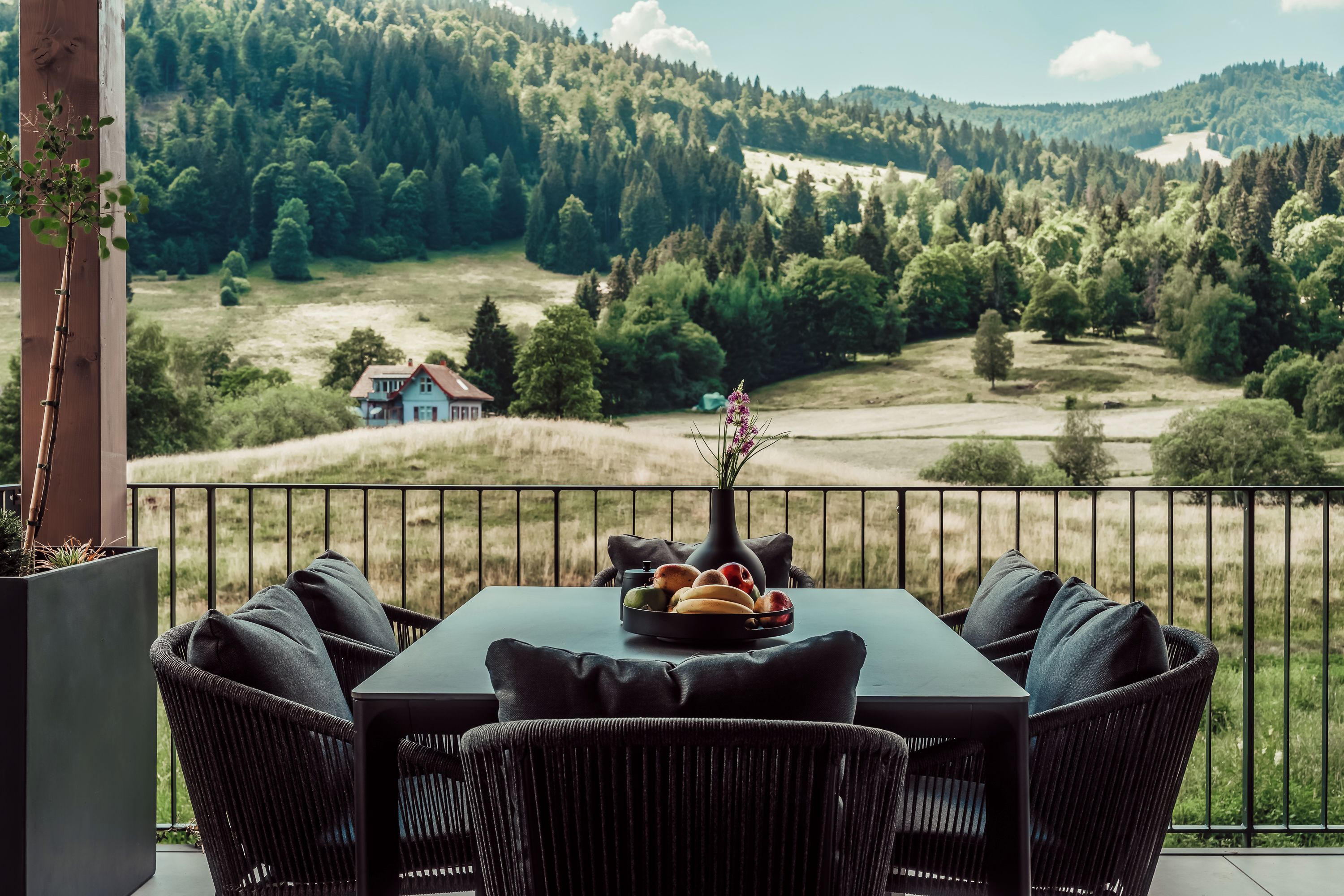 Un balcon avec une table et des chaises, un vase rempli de fleurs et une vue sur une vallée boisée.