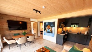 Un salon moderne avec un plafond en bois, un téléviseur à écran plat, un coin repas, une cuisine dotée de meubles sombres et un tapis vert.